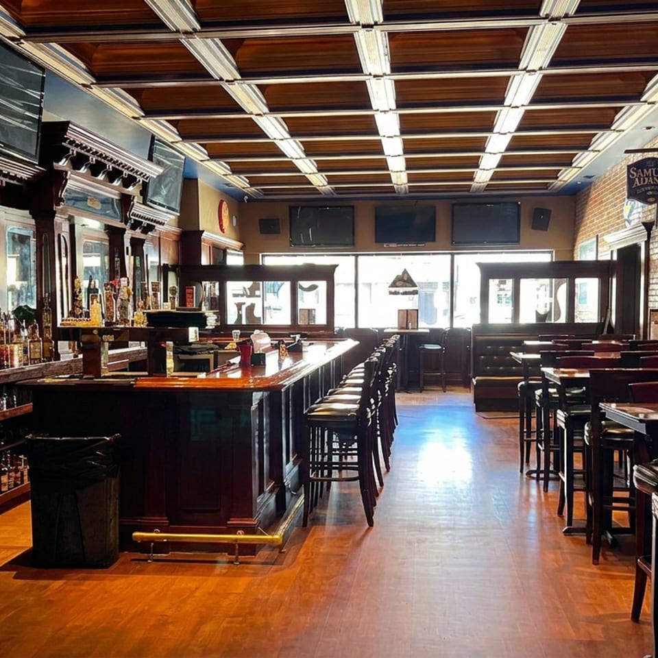 Empty upscale bar and restaurant interior with a polished wooden bar, liquor shelves, high-top tables, brick walls, and glowing ceiling lights.