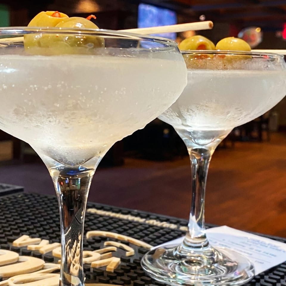 Two frosty cocktails in coupe glasses garnished with skewered green olives on a restaurant bar.