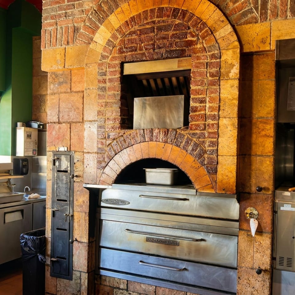 Restaurant kitchen featuring a brick arched wood-fired oven, stainless-steel cooking equipment, and green walls beside bright windows.