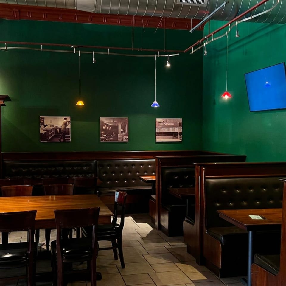 Empty restaurant dining room with dark green walls, wooden tables, leather booths, colorful pendant lights, and a restroom entrance.