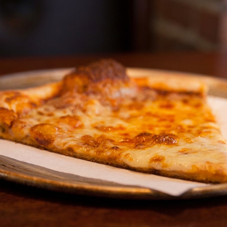 Large triangular slice of cheese pizza served on parchment paper atop a metal plate at a restaurant table.