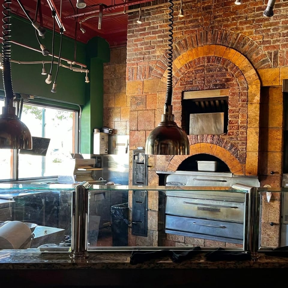 Restaurant pizza kitchen with brick ovens, hanging heat lamps, stainless-steel equipment, and a counter viewed through glass.