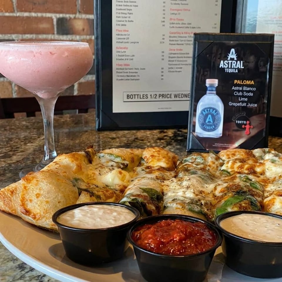 Stuffed pizza rolls with three dipping sauces beside a pink cocktail and Astral Tequila display on a restaurant table.