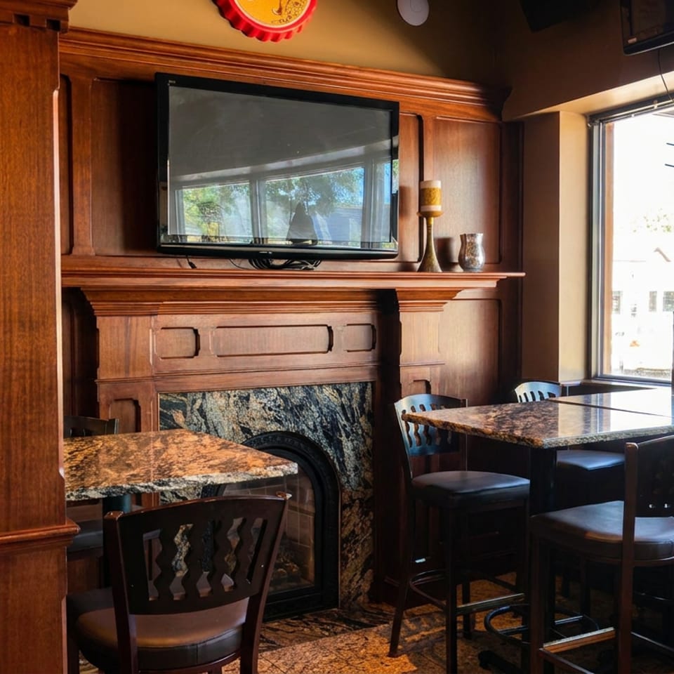 Warmly lit restaurant dining area with dark wood paneling, granite tables, leather chairs, fireplace, and street-facing windows.