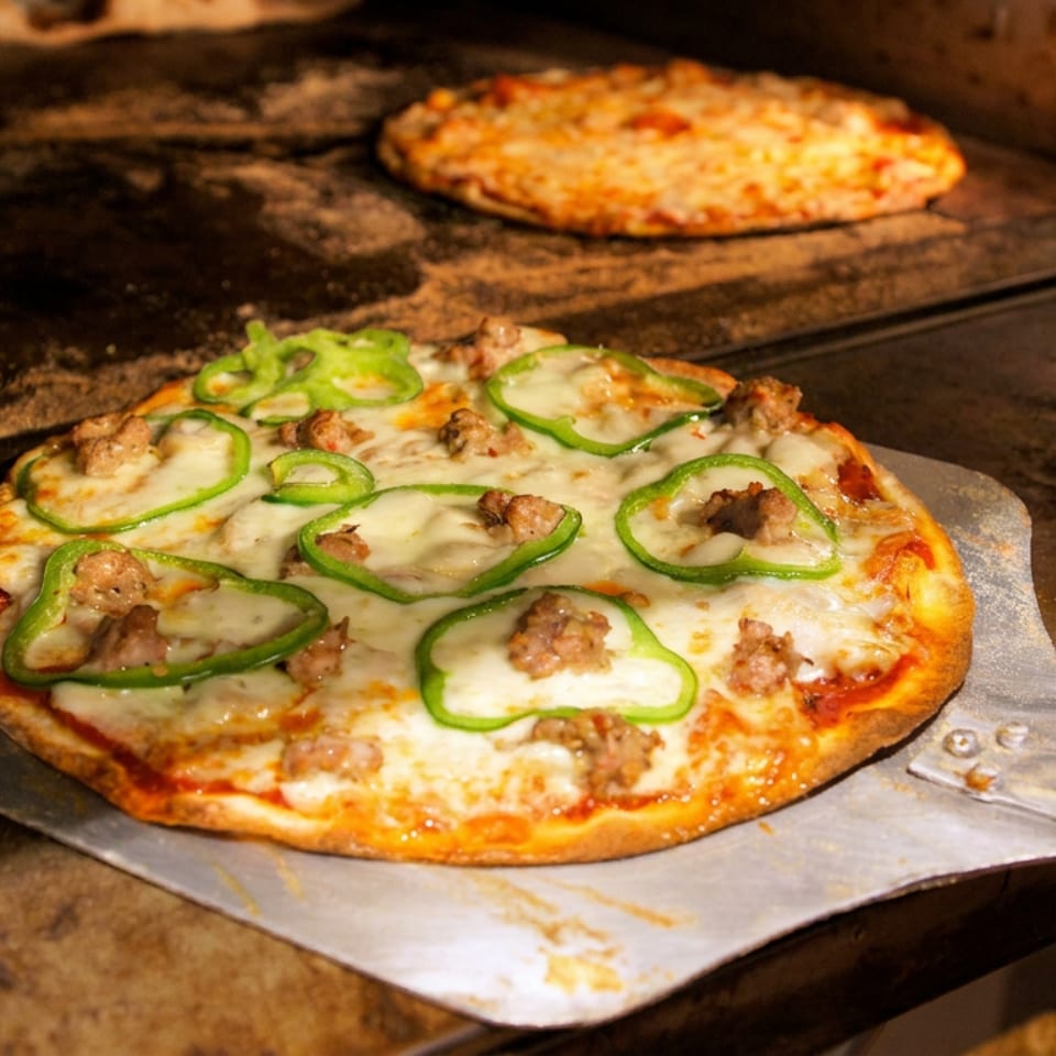 Freshly baked pizza topped with melted cheese, green peppers, and sausage rests on a metal peel inside a wood-fired oven.