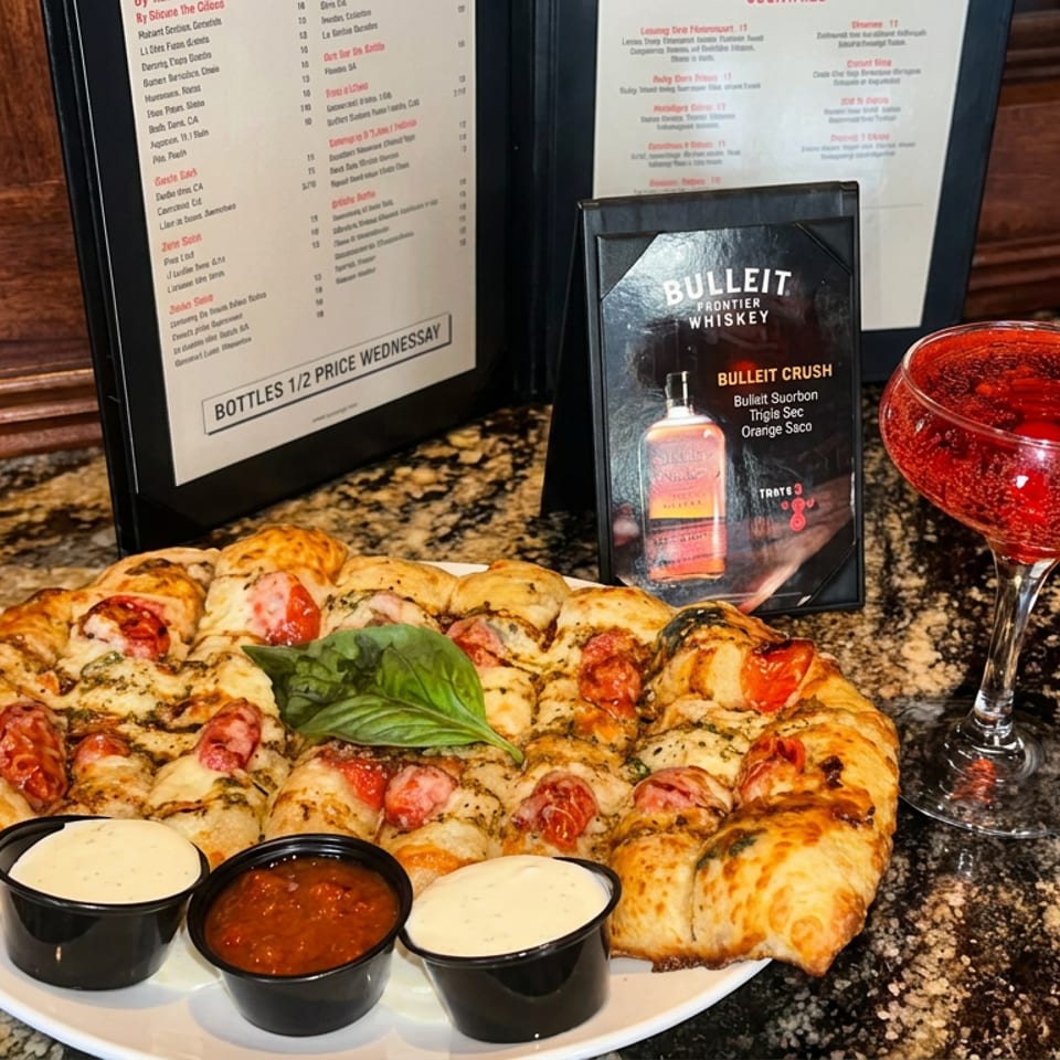 Pizza topped with tomatoes and basil sits beside dipping sauces and a red cocktail on a polished granite bar.